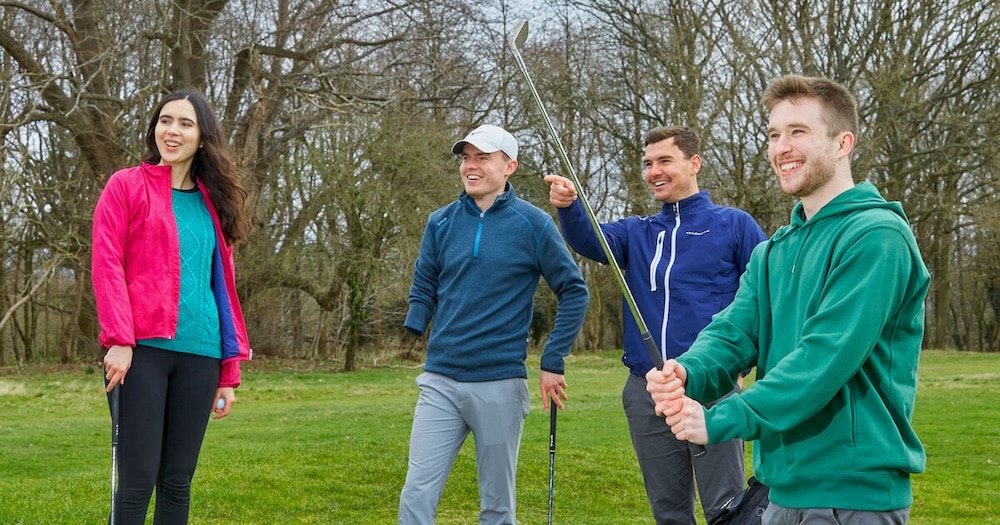 A family group of adults on the golf course with the male pointing at a golf ball in the distance whilst another adult male in a green jumper is swinging the golf club in the air.