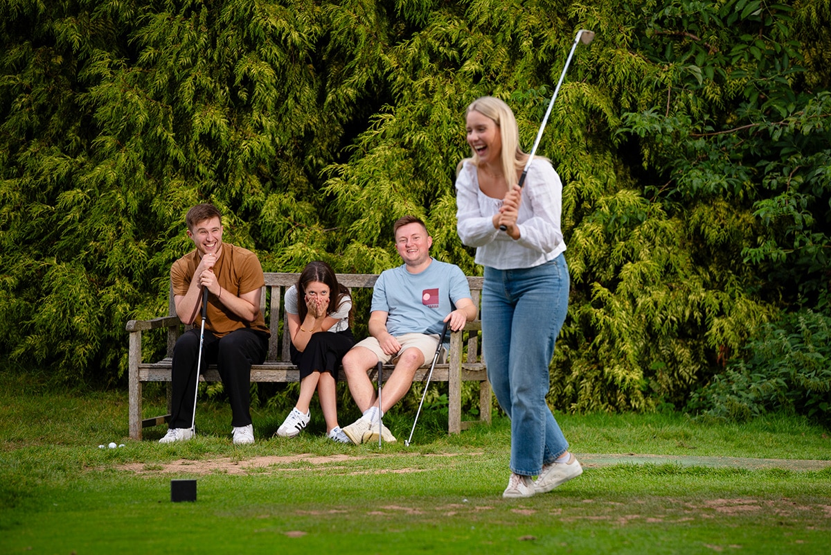 A group of friends on the golf course with 3 sat on a bench and an adult woman swinging her golf club towards the ball.