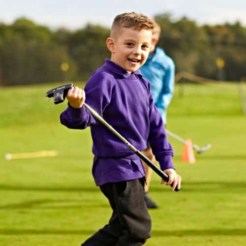 A young boy in a purple jumper is holding his junior golf club.