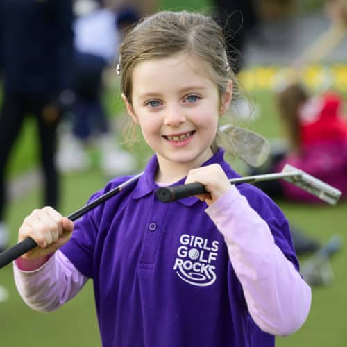 A young girl in a purple Girls Golf Rocks polo shirt is holding her junior club across her chest.