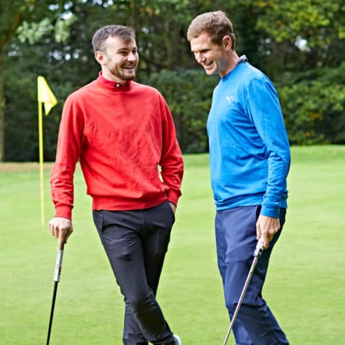 A male adult golfer in a red jumper shares a joke with a male golfer in a blue jumper on the golf course.