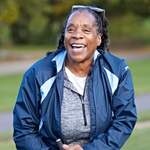 An adult female in a grey jumper and blue coat is laughing on the golf course.